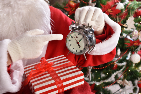 santa claus sitting with gifts in front of christmas tree  points his finger at alarm clockの写真素材