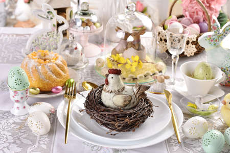 Easter table decoration with hen figurine in the nest on the plate, ring cake and decors in glass...