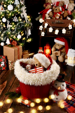 Teddy bear in Santa Claus cap with gift box in the basket in the room with Christmas tree and decorationsの写真素材
