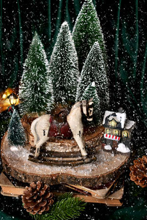 Christmas decoration with winter scene on a slice of wood with rocking horse, house and miniature Christmas trees and falling snowの写真素材