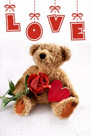Valentines card with cute teddy bear holding red rose and a heart sitting on white background with hanging inscription LOVEの写真素材