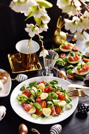 Easter breakfast with fresh vegetable salad with prosciutto ham, cucumber, avocado, cherry tomatoes, lettuce and quail eggs on black table with white and golden decorsの写真素材