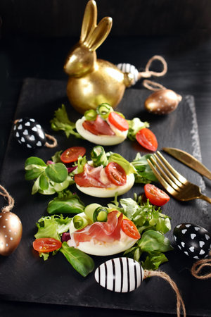 Easter breakfast with egg halves with prosciutto ham, cherry tomato, cucumber, avocado, mayonnaise and lettuce on black boardの写真素材