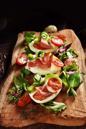 Egg halves with prosciutto ham, cherry tomato, cucumber, avocado, mayonnaise and lettuce on rustic wooden boardの写真素材