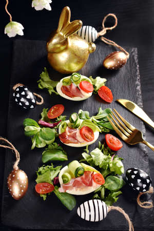 Top view of Easter breakfast with egg halves with prosciutto ham, cherry tomato, cucumber, avocado, mayonnaise and lettuce on black boardの写真素材