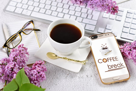 A cup of coffee, smartphone with "coffee break" display and white keyboard on the desk with lilac blossomsの写真素材