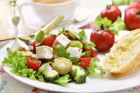 Fresh mediterraneanstyle salad with feta cheese, vegetables and bread on white plateの写真素材
