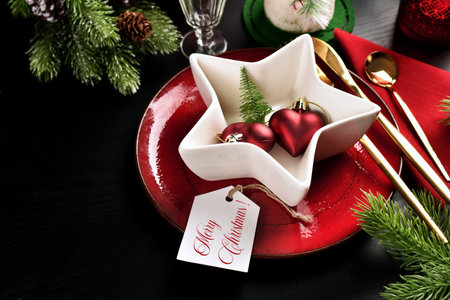 Christmas table setting with red plate and white star shaped bowl, cutlery and fir branches on black...