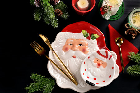 Christmas table setting with Santa Claus head shaped plate and bowl with cutlery, candles and fir branches on black surfaceの写真素材