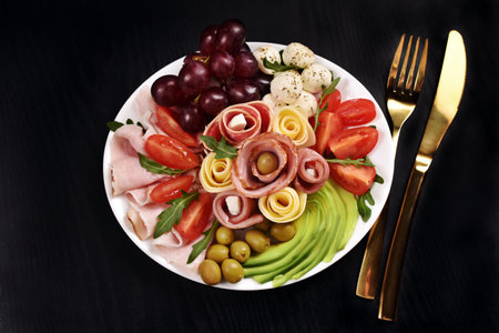 Top view of a platter with assortment of sliced ham, salami, cheese, avocado, mozzarella, grape with olives and tomatoes on black backgroundの写真素材