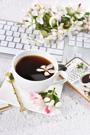 Cup of coffee, smartphone and computer keyboard in white on the desk in office or home with spring blossomsの写真素材
