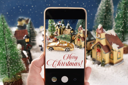 A hand holding the smartphone to take photos of miniature decoration of small town full of snow on Christmas night with wishesの写真素材