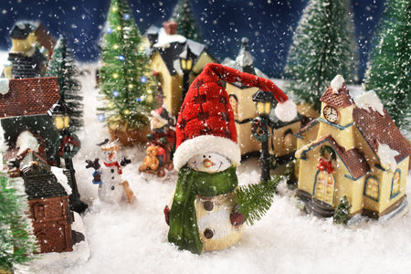 A cute snowman in Santa cap figurine standing in the middle of the street with houses in small town on snowy Christmas nightの写真素材