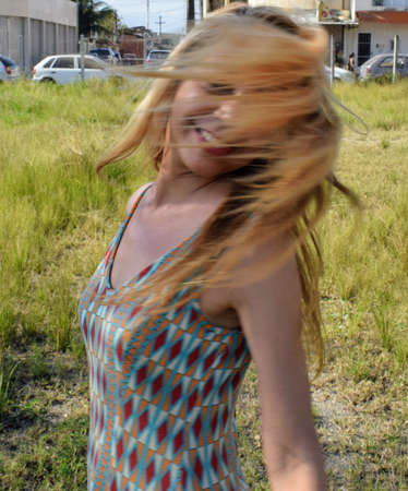 A young woman opens her arms, smiles and waves her blond hair that partially covers her face, in a wide grassy spot. In the background are seen houses and cars.の写真素材
