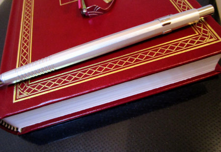 Side view of a silver mechanical pencil and a pair of eyeglasses over a red hardcover book with gold...