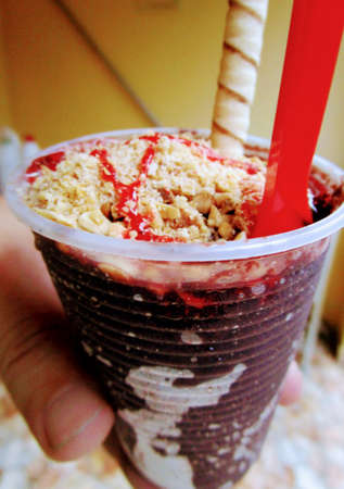 Hand holding a glass of iced acai berry, with nuts and various toppings, waffle and red plastic spoon, to eat for dessert.の写真素材
