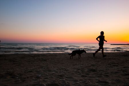 Beautiful young woman running on a beach at sunset with her dogの写真素材