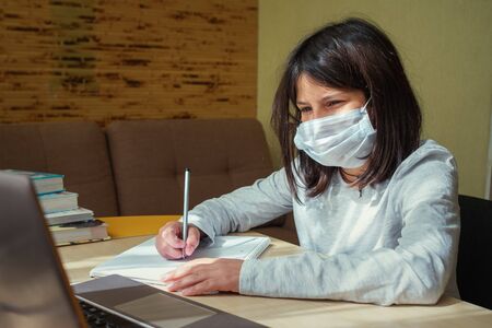 A schoolgirl with a medical mask on her face watches an online lesson and takes notes. The concept of home schooling during quarantine.の写真素材