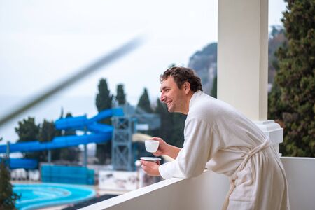 A happy smiling man in a white bathrobe is standing with a cup of morning tea or coffee standing on the balcony of his room against the backdrop of the water slides of the sea..の写真素材