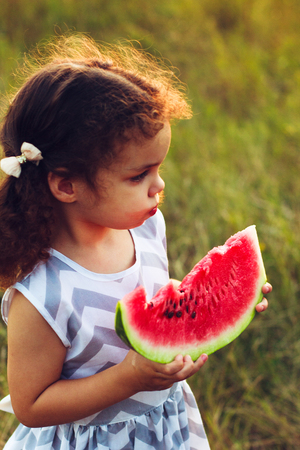 Funny portrait of an incredibly beautiful curly-haired little girl eating watermelon, healthy fruit snack, adorable toddler child with curly hair playing in a sunny park on a hot summer day. portraitの写真素材