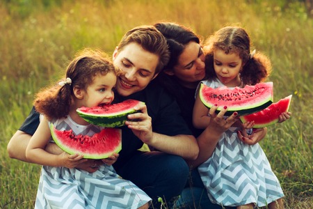 Cheerful positive family of four having a picnic and eating watermelon outdoors in a sunny weather. Curly beautiful twins sisters eating a snack lunch. Vegetarian Concept. Happy people.の写真素材