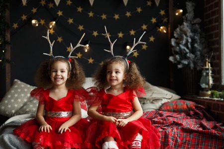 Cute curly twins girl sitting on the bed in decorated xmas room, on Christmas evening 2018 near New Year fir tree with lights and garlands. Sisters wearing red dresses and deer corns hoopの写真素材