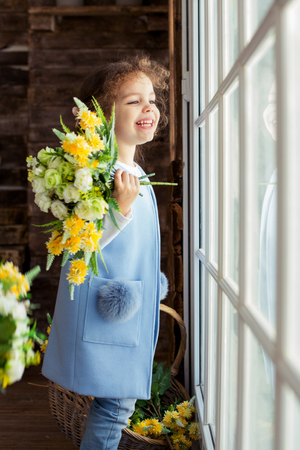 Adorable little child girl sitting on the window and holding flowers. Girl looks at the blue, pure sky and rejoices to spring and sun.の写真素材