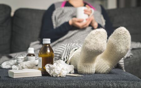 Cold medicine and sick woman drinking hot beverage to get well from flu, fever and virus. Dirty paper towels and tissues on table. Ill person wearing warm woolen stocking socks in winter.の写真素材