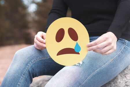 Crying paper emoticon in hand. Depressed woman holding printed sad smiley face and sitting on a rock. Modern communication and smiley icon on cardboard. Sadness, depression and emotions concept.の写真素材