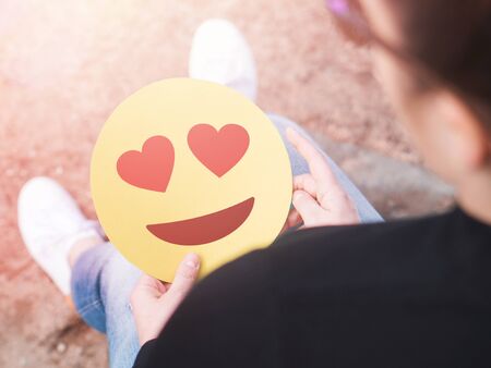 Heart emoticon in hand. Smitten young woman holding printed paper or cardboard love smiley face. Happy Valentine's day or anniversary theme. Creative communication. Being in love or having a crush concept.の写真素材