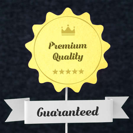 Premium quality guaranteed badge cut from cardboard and paper on a wooden stick. Yellow banner and ribbon for business website to promise customer the best product or service. Seal on dark background.の写真素材