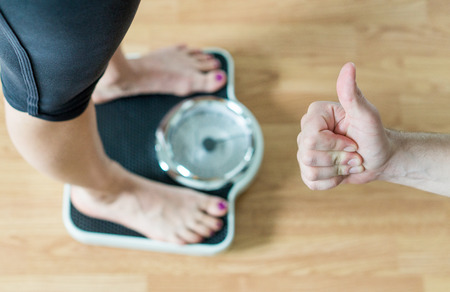 Personal trainer or coach giving thumbs up to a person standing on scale. Success and great...