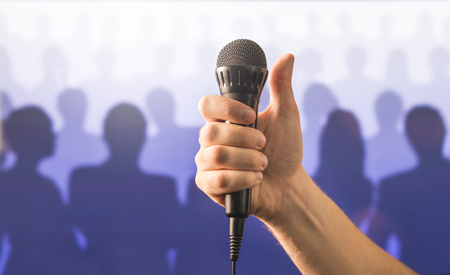 Hand holding microphone and showing thumbs up in front of a crowd of silhouette people. Public speaking and giving speech concept. Singing well live to mic, good karaoke or successful talent show.の写真素材