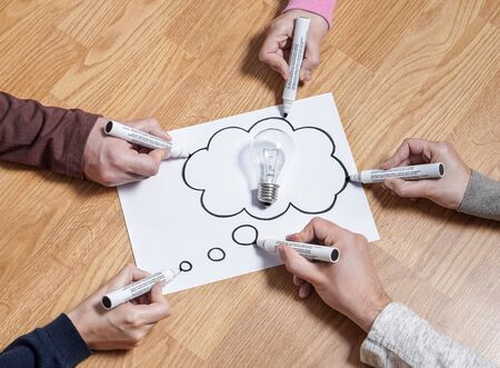 Thinking speech bubble balloon with light bulb. Brainstorming new ideas together for marketing plan or school project. Teamwork, synergy and education. Group of people drawing to same paper.の写真素材