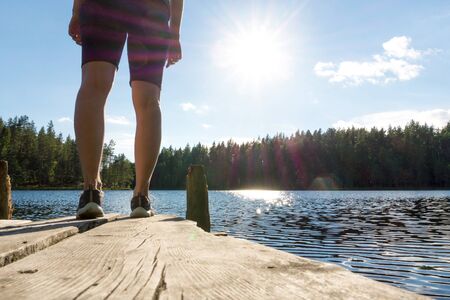 Young woman standing on an old wooden dock and pier at a lake at summer in Finland. Sun shining in blue sky. Traditional Finnish nature view.の写真素材