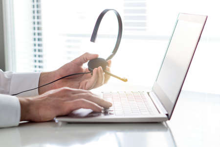 Help desk, support hotline or call center concept. Video call conference or telemarketing. Customer service person holding headset in hand and typing chat with laptop.の写真素材