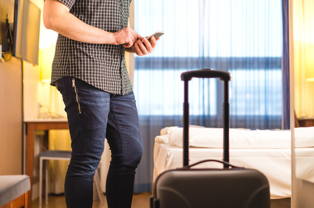 Man using smartphone in hotel room with baggage and suitcase. Tourist with mobile phone in holiday rental apartment. Calling taxi cab or doing online check-in. Travel and smart technology concept.の写真素材