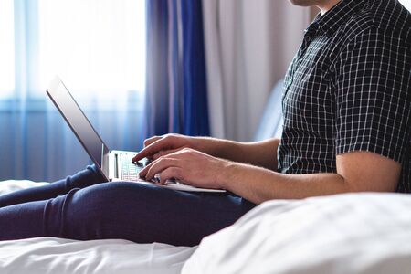 Man using laptop in hotel room or home bedroom. Guy lying on bed and typing with notebook computer. Person in the morning with modern device, reading emails and sending messages. Telecommuting conceptの写真素材