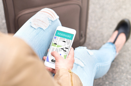 Rideshare app on smartphone. Young woman using ride sharing application and service with mobile phone while traveling. Ordering taxi or carpool ride. Sitting with baggage and suitcase.の写真素材