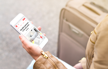 Woman search holiday apartments and rooms online with mobile phone. Vacation home rental website or application on smartphone screen. Traveler sitting next to suitcase, baggage or luggage.の写真素材