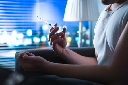 Drug addict holding heroin needle in hand. Young man with hypodermic syringe. Dramatic addiction, overdose and narcotic abuse concept.の写真素材