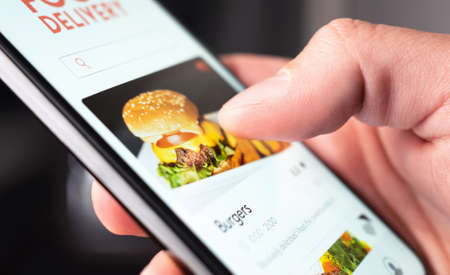 Food delivery app order with phone. Online mobile service for take away burger and pizza. Hungry man reading restaurant menu, website and reviews with smartphone. Takeout or fast courier deliver.の写真素材