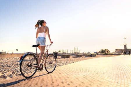Bicycle fun on beach promenade. Happy woman riding bike on sunny summer boulevard. Seaside waterfront street for cycling. Stylish cyclist lady. Cool carefree feeling at sunset. Summertime freedom.の写真素材