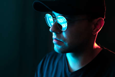 Man with neon light on glasses and face. Blue and red color from city billboard in dark studio portrait. Reflection in sunglasses. Black background in dramatic urban night portrait. Cyber technology.の写真素材