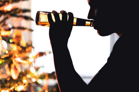 Christmas and alcohol problem. Lonely man drinking beer. Xmas tree. Alcoholism alone during holiday. Sip from bottle of lager. Sad guy with depression and addiction. Silhouette of wasted alcoholic.の写真素材