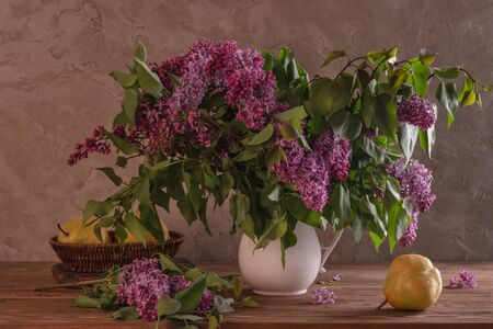 Spring still life lilac with a white vase with fresh pears on a wooden table against the background of an old wallの写真素材