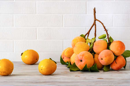 A branch of ripe persimmons on a wooden table against a background of a white brick wall