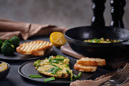 Omelet with broccoli and green onions on a black plate and frying pan on a black backgroundの写真素材