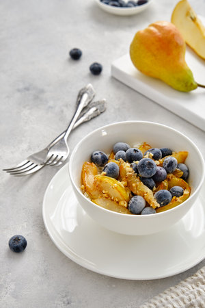 Cottage cheese with pieces of pears, nuts and blueberries in a white cup on a light-gray background. Delicious and healthy breakfastの写真素材