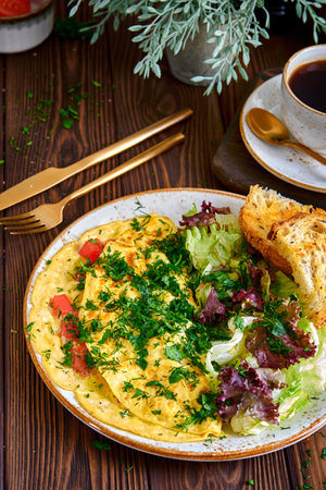 Omelette with tomatoes and herbs on a beige plate with a cup of black coffee on a wooden table close-upの写真素材